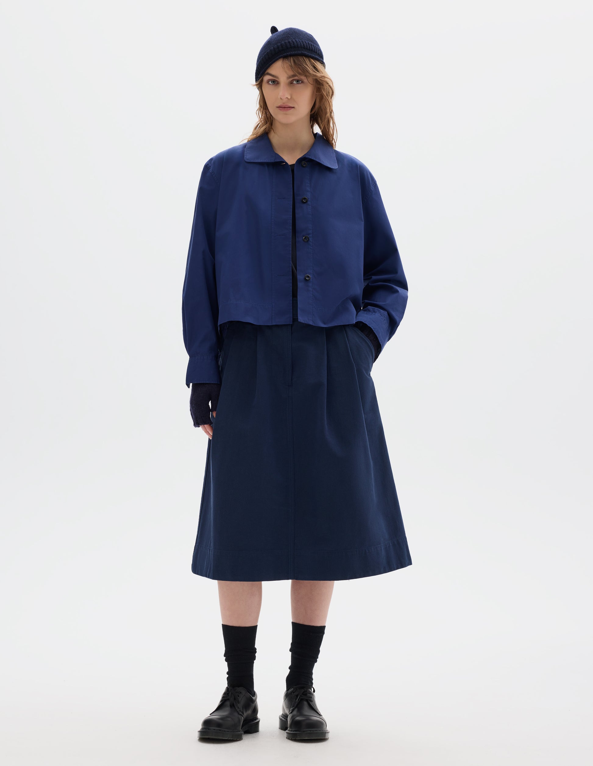 Full body image, front view of model wearing an asymmetric blue cotton shirt, with collar with a softly rounded point and dark buttons. Styled with a dark navy wool bobble hat, fingerless gloves, blue pleated skirt, black socks and leather shoes.