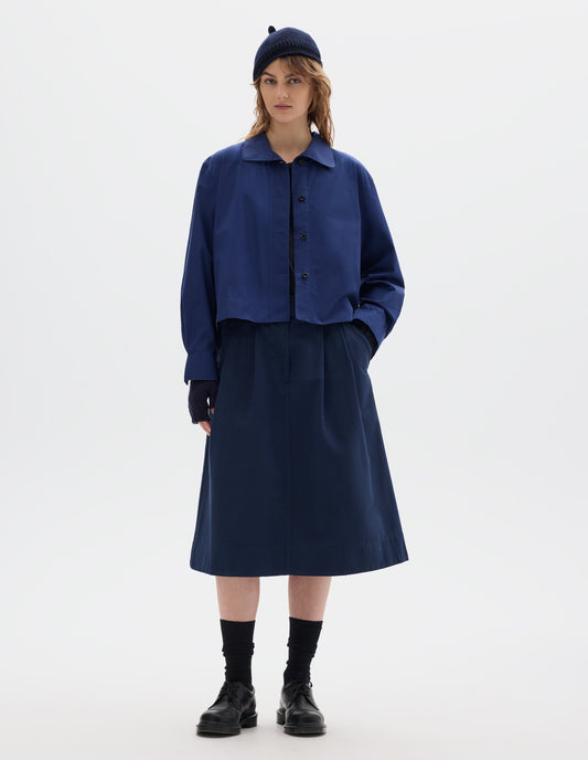 Full body image, front view of model wearing an asymmetric blue cotton shirt, with collar with a softly rounded point and dark buttons. Styled with a dark navy wool bobble hat, fingerless gloves, blue pleated skirt, black socks and leather shoes.
