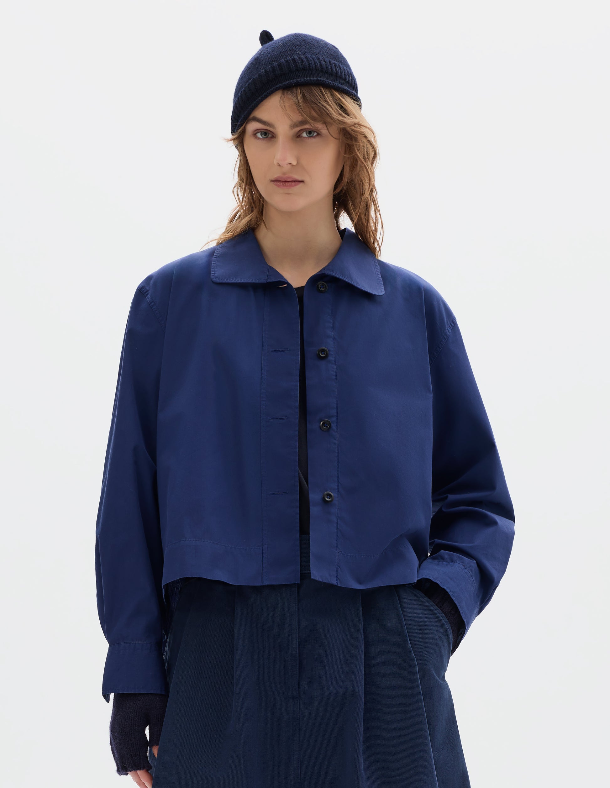 Cropped body image, front view of model wearing an asymmetric blue cotton shirt, with collar with a softly rounded point and dark buttons. Styled with a dark navy wool bobble hat, fingerless gloves and blue pleated skirt.