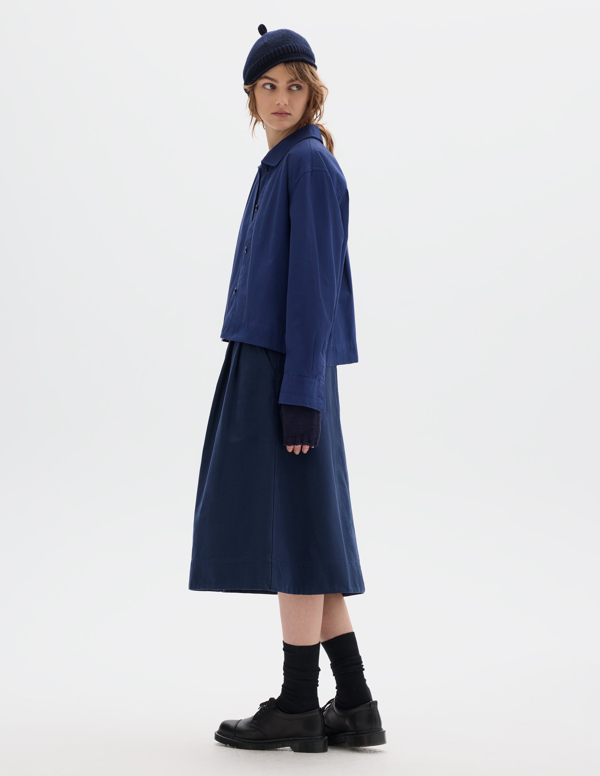 Full body image, front side view of model wearing an asymmetric collar blue cotton shirt, with a boxy cropped cut. Styled with a dark navy wool bobble hat, fingerless gloves, blue pleated A line skirt, black socks and leather shoes.