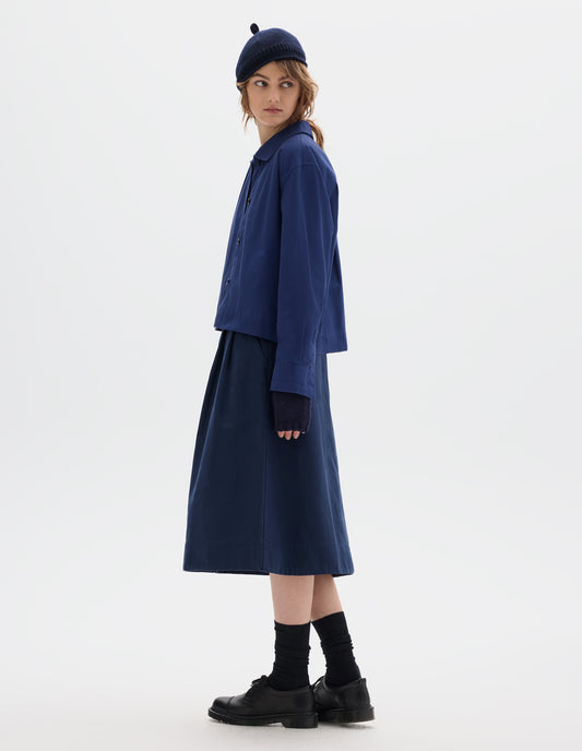Full body image, front side view of model wearing an asymmetric collar blue cotton shirt, with a boxy cropped cut. Styled with a dark navy wool bobble hat, fingerless gloves, blue pleated A line skirt, black socks and leather shoes.
