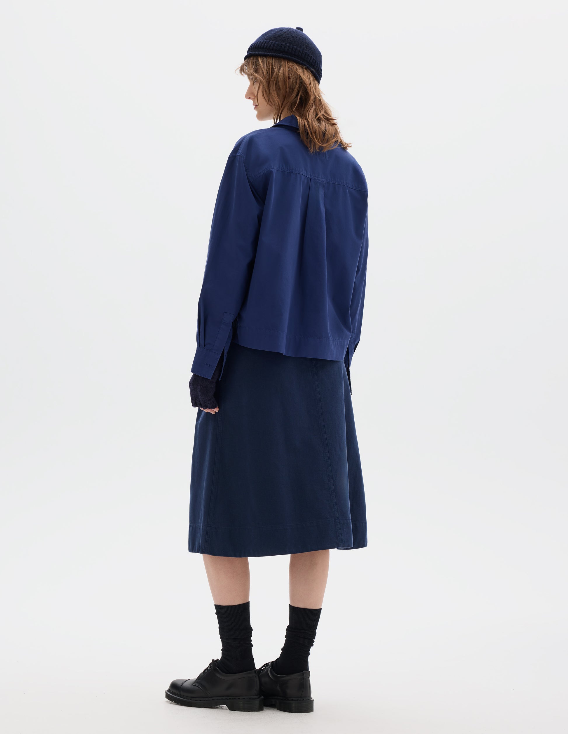 Back body image, front side view of model wearing a blue cotton shirt, with a boxy cropped cut, box pleat on back yoke. Styled with a dark navy wool bobble hat, fingerless gloves, blue A line skirt, black socks and leather shoes.