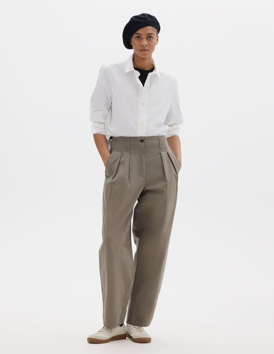 Full body image, front view of model wearing brown cotton trousers, with front pleats, a brown button with belt loops and side pockets. Styled with black t shirt, white shirt, black hat and white shoes.