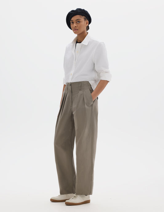 Full body image, side view of model wearing brown cotton trousers, with front pleats, a brown button with belt loops and side pockets. Styled with black t shirt, white shirt, black hat and white shoes.