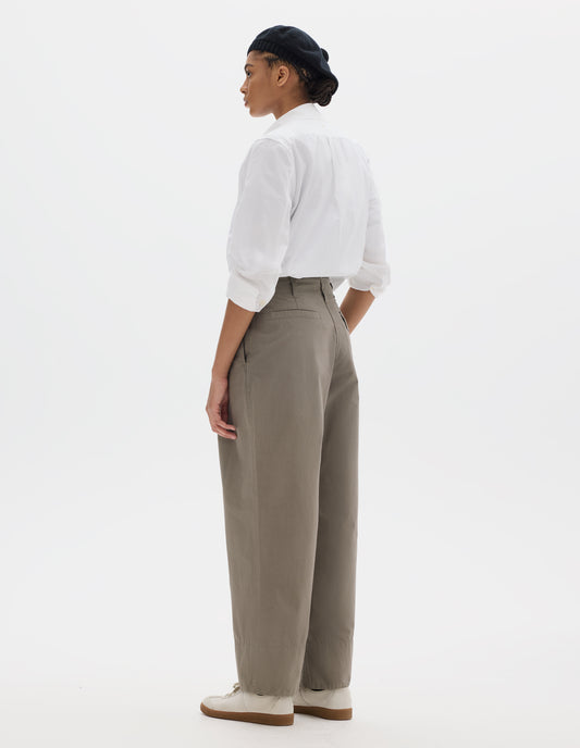 Full body image, back view of model wearing brown cotton trousers, with front pleats, a brown button with belt loops and side pockets. Styled with black t shirt, white shirt, black hat and white shoes.