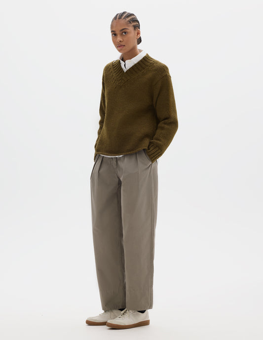 Full body image, front view of model wearing a brown green wool chunky wool sweater with a deep ribbed v neck, relaxed sleeves with rib cuffs and cast-off hem. Styled over a white shirt and with a greyish beige pleated trousers and off white suede trainers.