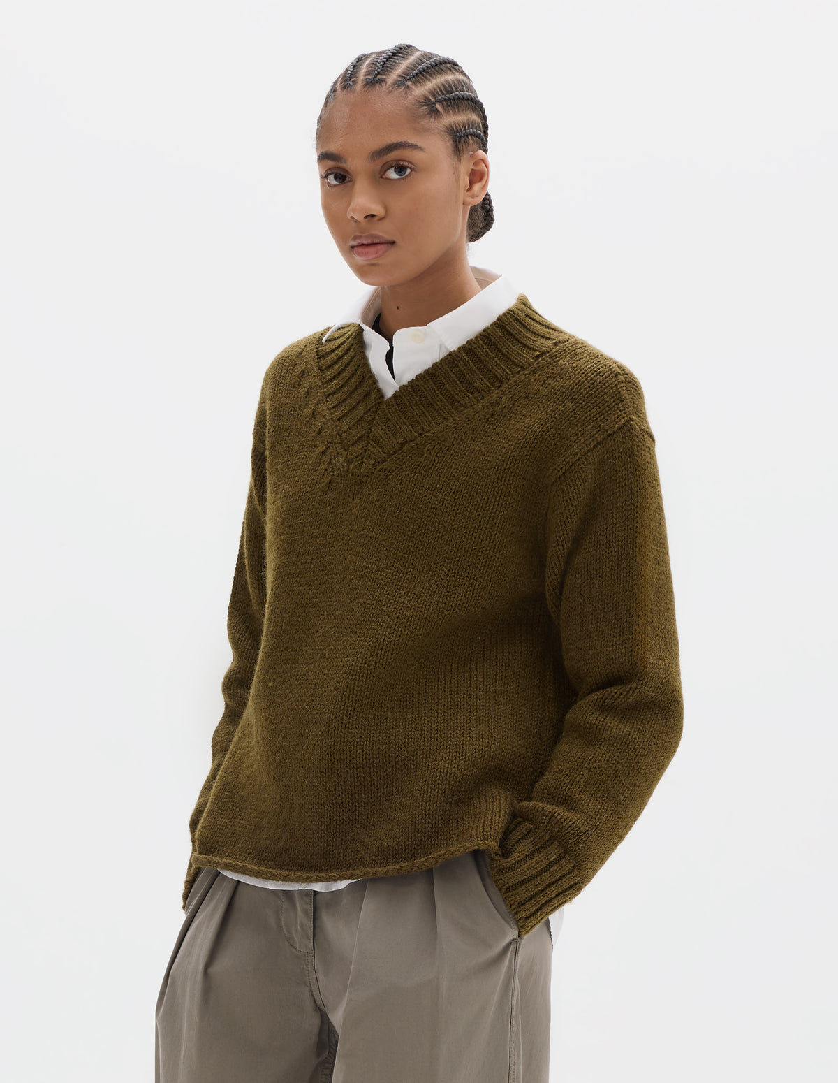 Cropped body image, front view of model wearing a brown green wool chunky wool sweater with a deep ribbed v neck, relaxed sleeves with rib cuffs and cast-off hem. Styled over a white shirt and with a greyish beige pleated trousers.