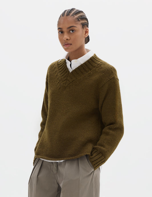 Cropped body image, front view of model wearing a brown green wool chunky wool sweater with a deep ribbed v neck, relaxed sleeves with rib cuffs and cast-off hem. Styled over a white shirt and with a greyish beige pleated trousers.
