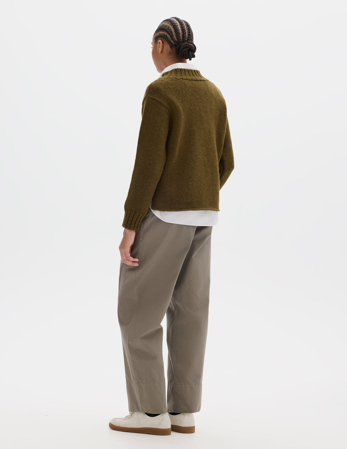 Full body image, back view of model wearing a brown green wool chunky wool sweater with a deep ribbed neck, relaxed sleeves with rib cuffs and cast-off hem. Styled over a white shirt and with a greyish beige trousers with a deep hem and off white suede trainers.
