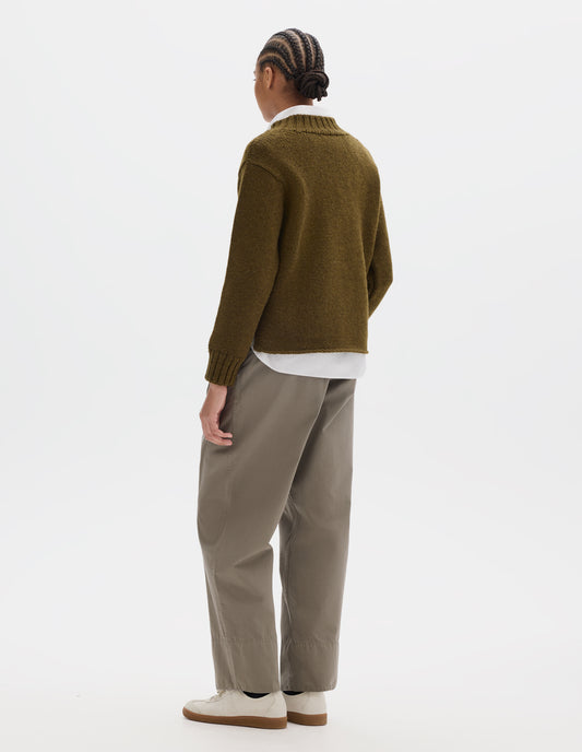 Full body image, back view of model wearing a brown green wool chunky wool sweater with a deep ribbed neck, relaxed sleeves with rib cuffs and cast-off hem. Styled over a white shirt and with a greyish beige trousers with a deep hem and off white suede trainers.