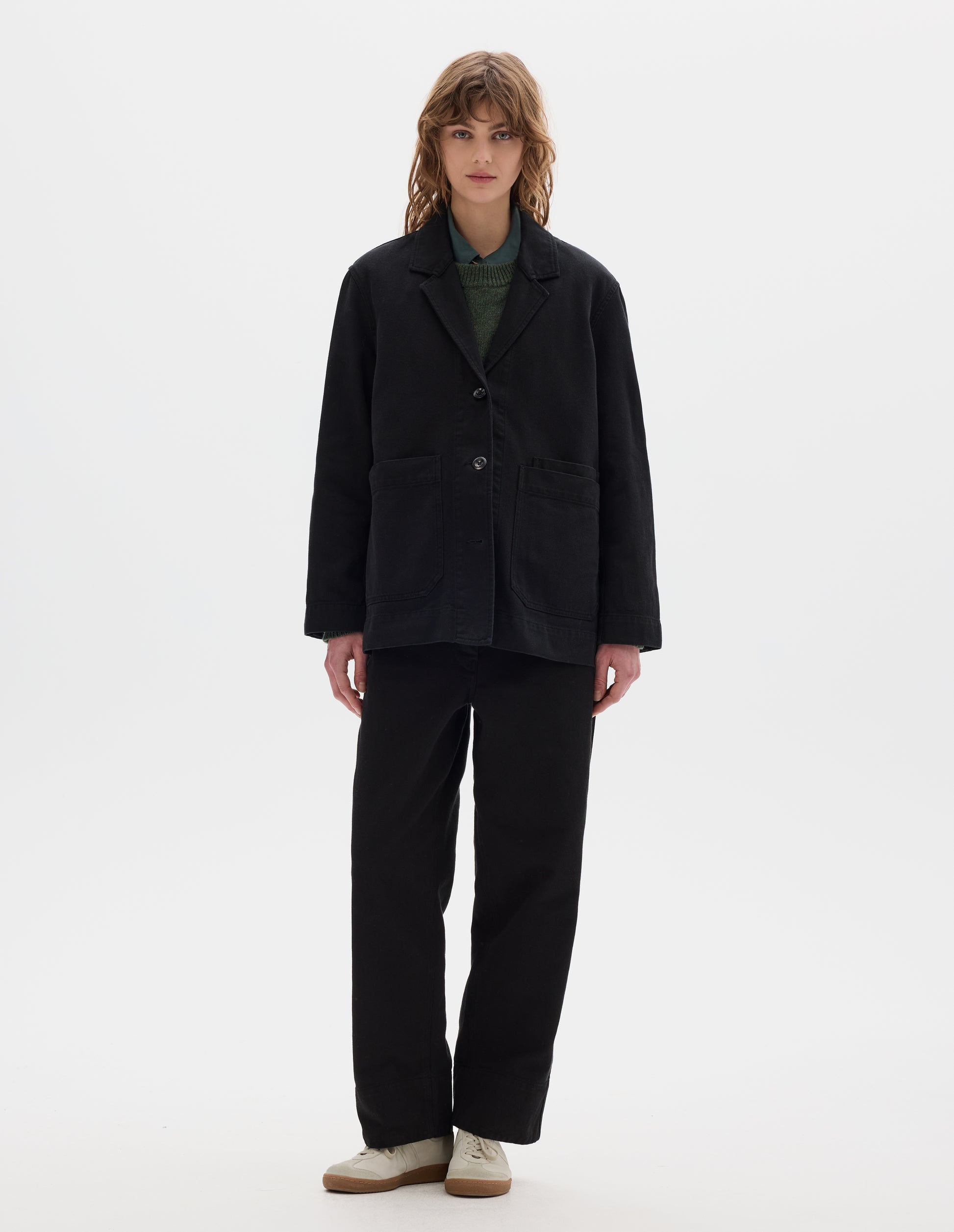 Full body image, front view of model wearing a black cotton blazer with a low breakpoint and three button closure, a wide placket, big front patch pocket with a double pocket detail and grown-on cuffs. Styled with blue shirt, green jumper, black trousers and white suede trainers.