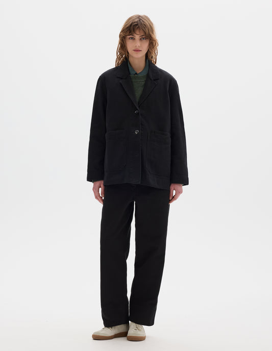 Full body image, front view of model wearing a black cotton blazer with a low breakpoint and three button closure, a wide placket, big front patch pocket with a double pocket detail and grown-on cuffs. Styled with blue shirt, green jumper, black trousers and white suede trainers.