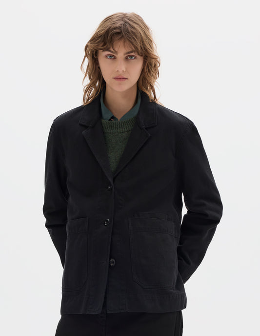 Cropped body image, front view model wearing a black cotton blazer with a low breakpoint and three button closure, a wide placket, big front patch pocket with a double pocket detail and grown-on cuffs. Styled with blue shirt, green jumper, and black trousers.