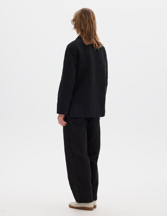 Full body image, back view of model wearing a black cotton blazer with a low breakpoint and three button closure, a wide placket, big front patch pocket with a double pocket detail and grown-on cuffs. Styled with blue shirt, green jumper, black trousers and white suede trainers.
