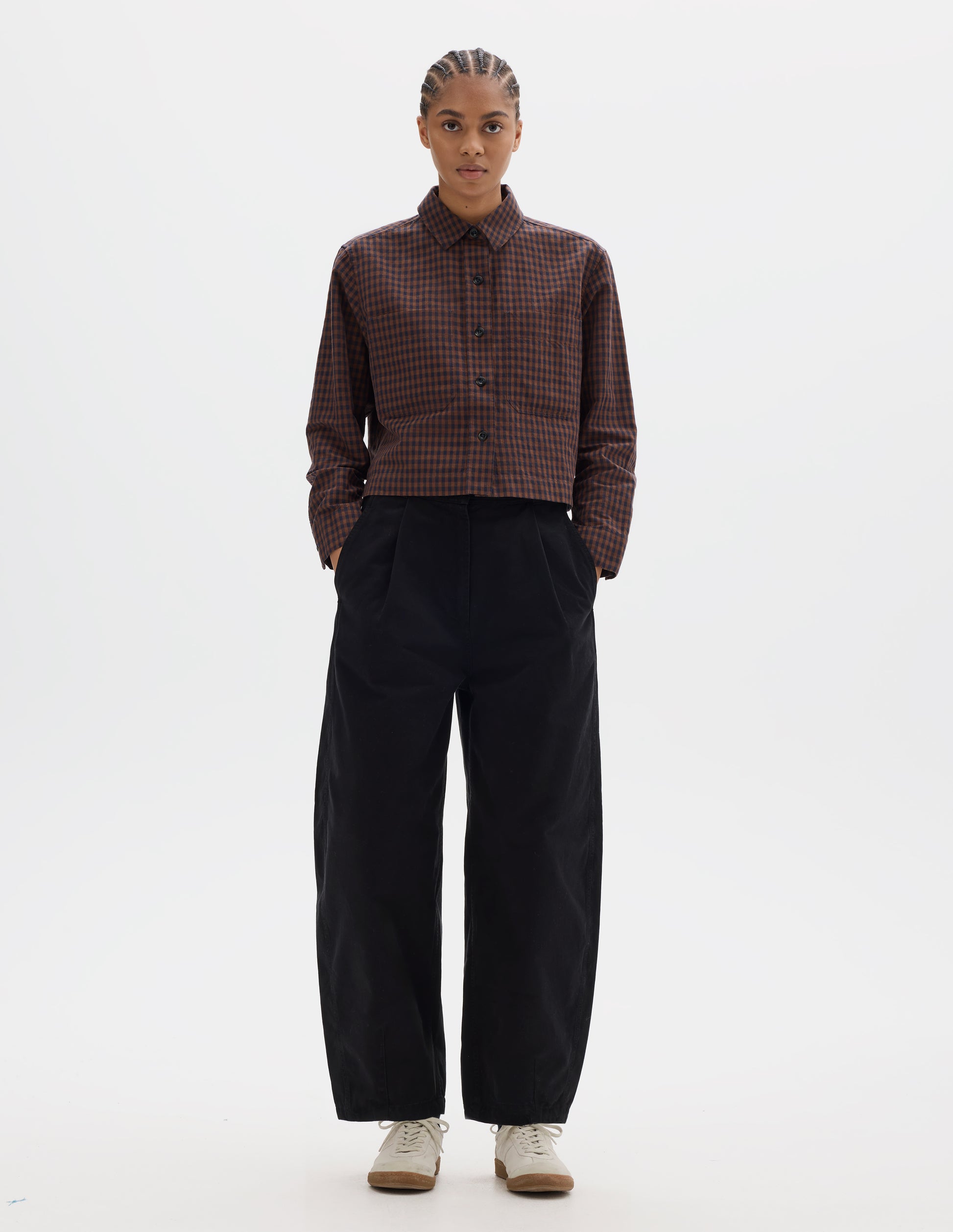 Full body image, front view of model wearing brown and dark blue check cotton linen shirt, with patch chest pockets and dark buttons. Styled with black pleated trousers and white gum sole trainers.