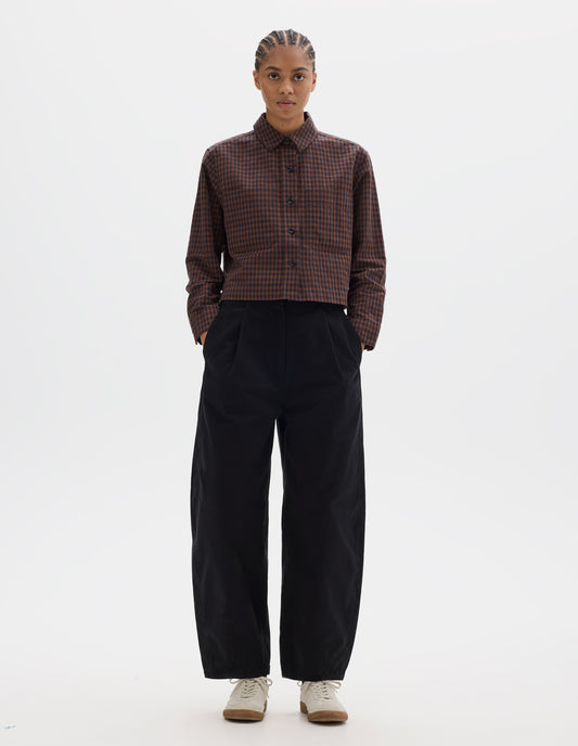 Full body image, front view of model wearing brown and dark blue check cotton linen shirt, with patch chest pockets and dark buttons. Styled with black pleated trousers and white gum sole trainers.
