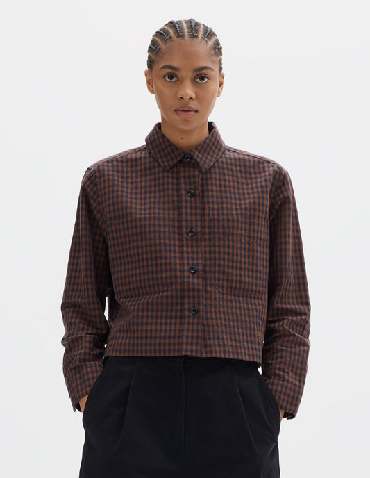 Cropped body image, front view of model wearing brown and dark blue check cotton linen shirt, with patch chest pockets and dark buttons. Styled with black pleated trousers.