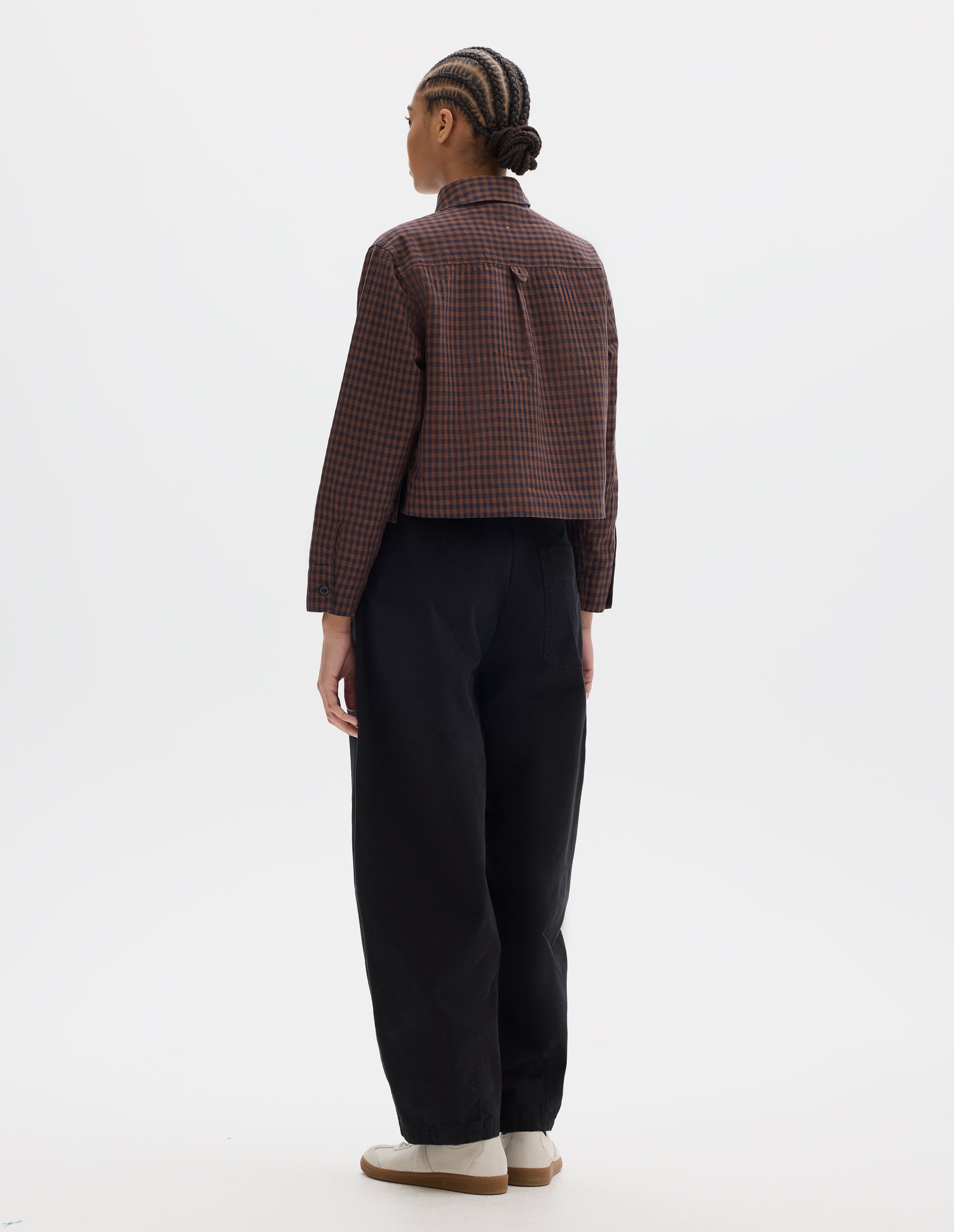 Full body image, back view of model wearing brown and dark blue check cotton linen shirt, with back yoke, box pleat, external hem loop detailing and button cuffs. Styled with black trousers and white trainers with gum sole.