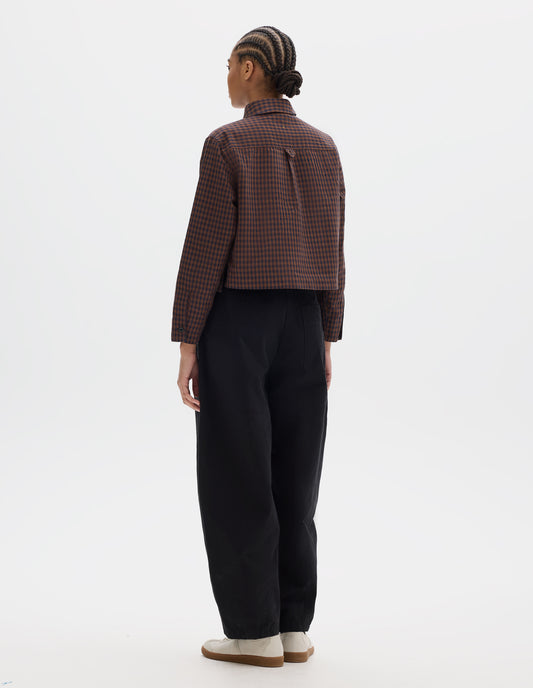 Full body image, back view of model wearing brown and dark blue check cotton linen shirt, with back yoke, box pleat, external hem loop detailing and button cuffs. Styled with black trousers and white trainers with gum sole.