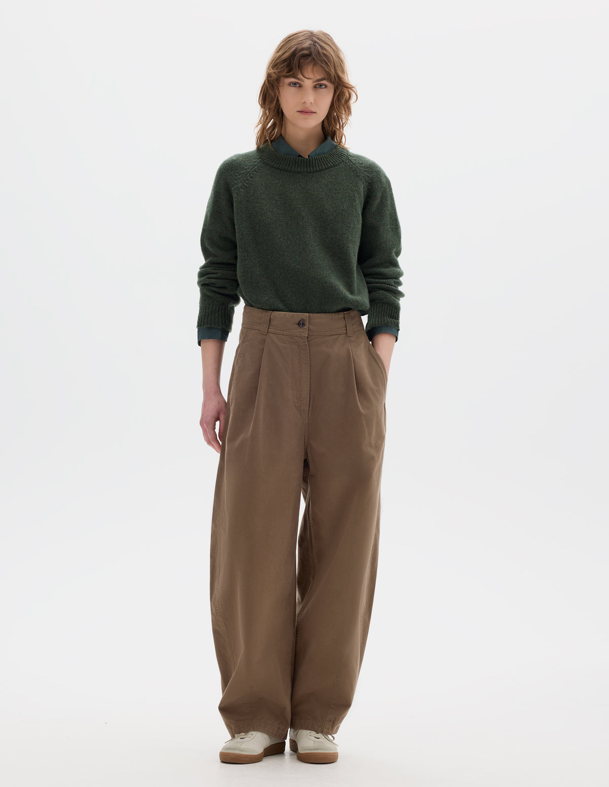 Full body image, front view of model wearing a light brown cotton trousers with a deep single front pleat, darts at the hem and side pockets. Styled with green wool crew neck sweater, worn over a greenish blue shirt and off white suede trainers.