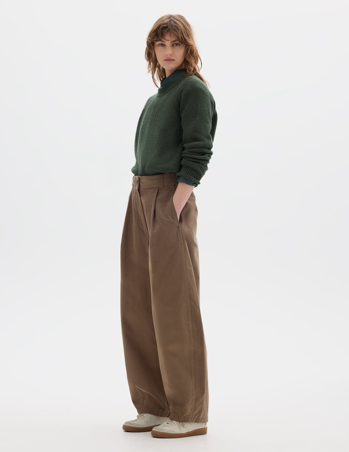 Full body image, alternative side front view of model wearing a light brown cotton trousers with a deep single front pleat, darts at the hem and side pockets. Styled with green wool crew neck sweater, sleeves rolled up, worn over a greenish blue shirt and off white suede trainers.