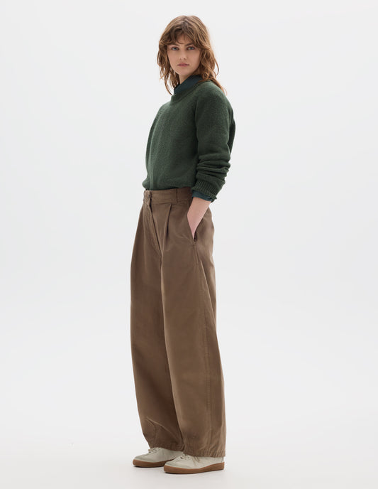 Full body image, alternative side front view of model wearing a light brown cotton trousers with a deep single front pleat, darts at the hem and side pockets. Styled with green wool crew neck sweater, sleeves rolled up, worn over a greenish blue shirt and off white suede trainers.