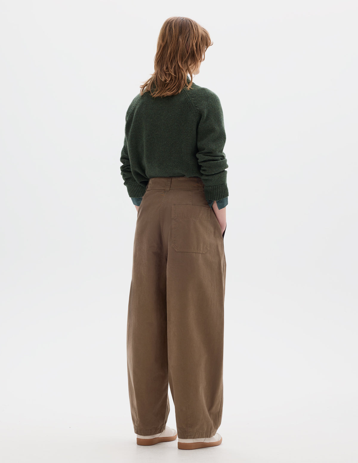 Full body image, back view of model wearing a light brown cotton trousers with a back patch pocket, darts at the hem and side pockets. Styled with green wool crew neck sweater, sleeves rolled up, worn over a greenish blue shirt and off white suede trainers.