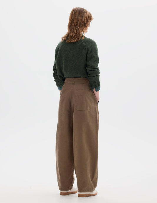Full body image, back view of model wearing a light brown cotton trousers with a back patch pocket, darts at the hem and side pockets. Styled with green wool crew neck sweater, sleeves rolled up, worn over a greenish blue shirt and off white suede trainers.