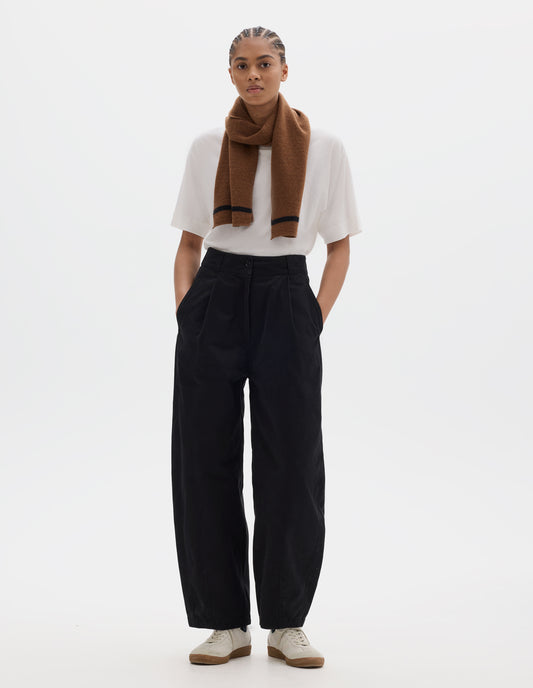 Full body image, front view of model wearing a black cotton trousers with a deep single front pleat, darts at the hem and side pockets. Styled with a white short sleeve t shirt, brown wool scarf with a black tip and off white suede trainers.