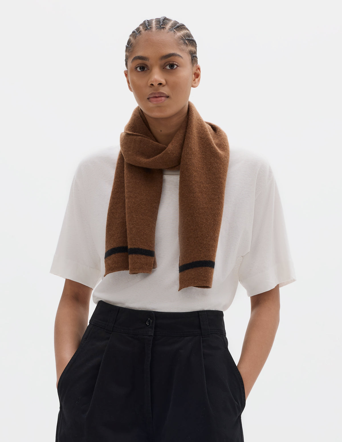 Cropped body image, front view of model wearing an orangey brown lambswool scarf with a black stripe, styled with a short sleeve white t shirt and black pleated trousers.