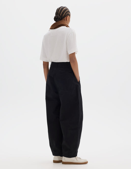 Full body image, back view of model wearing a black cotton trousers with darts at the hem, side pockets and a back patch pocket. Styled with a white short sleeve t shirt, brown wool scarf and off white suede trainers.