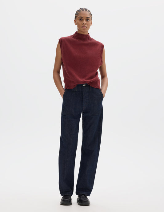 Full body image, front view of model wearing indigo blue denim twill jeans, with metal button fly, stitch-through waistband with belt loops, and stitch through pockets. Styled with red sleeveless high neck wool slipover and black leather shoes.