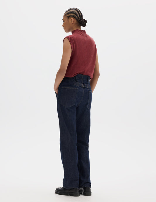 Full body image, back view of model wearing indigo blue denim twill jeans, with stitch-through waistband with belt loops, cinch back detail with metal buckle and patch back pockets. Styled with red sleeveless high neck wool slipover and black leather shoes.