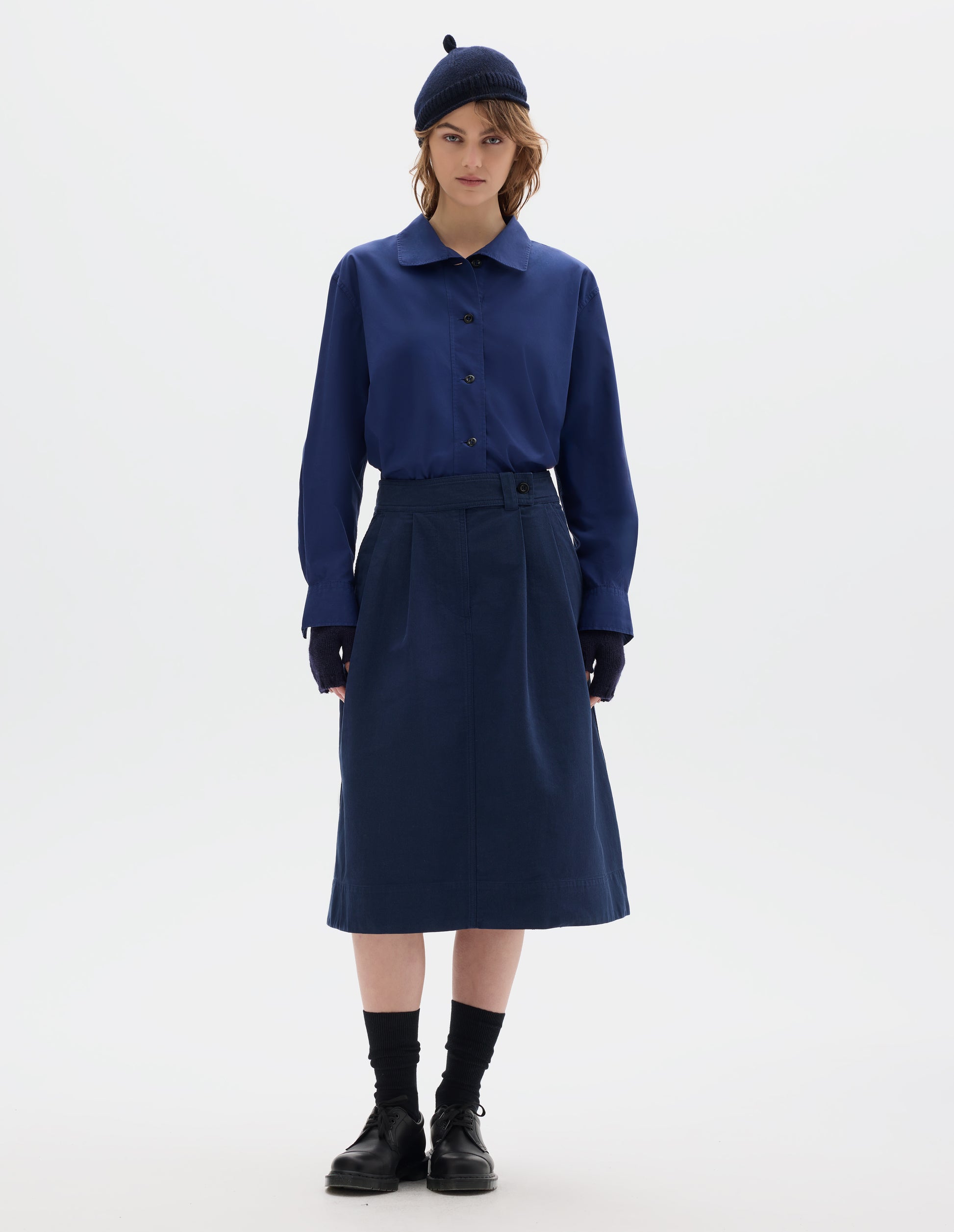 Full body image, front view of model wearing dark blue cotton skirt, with loop and button fastening, slant pockets and ticket pockets. Styled with blue shirt, navy hat and fingerless gloves, black socks and black shoes.