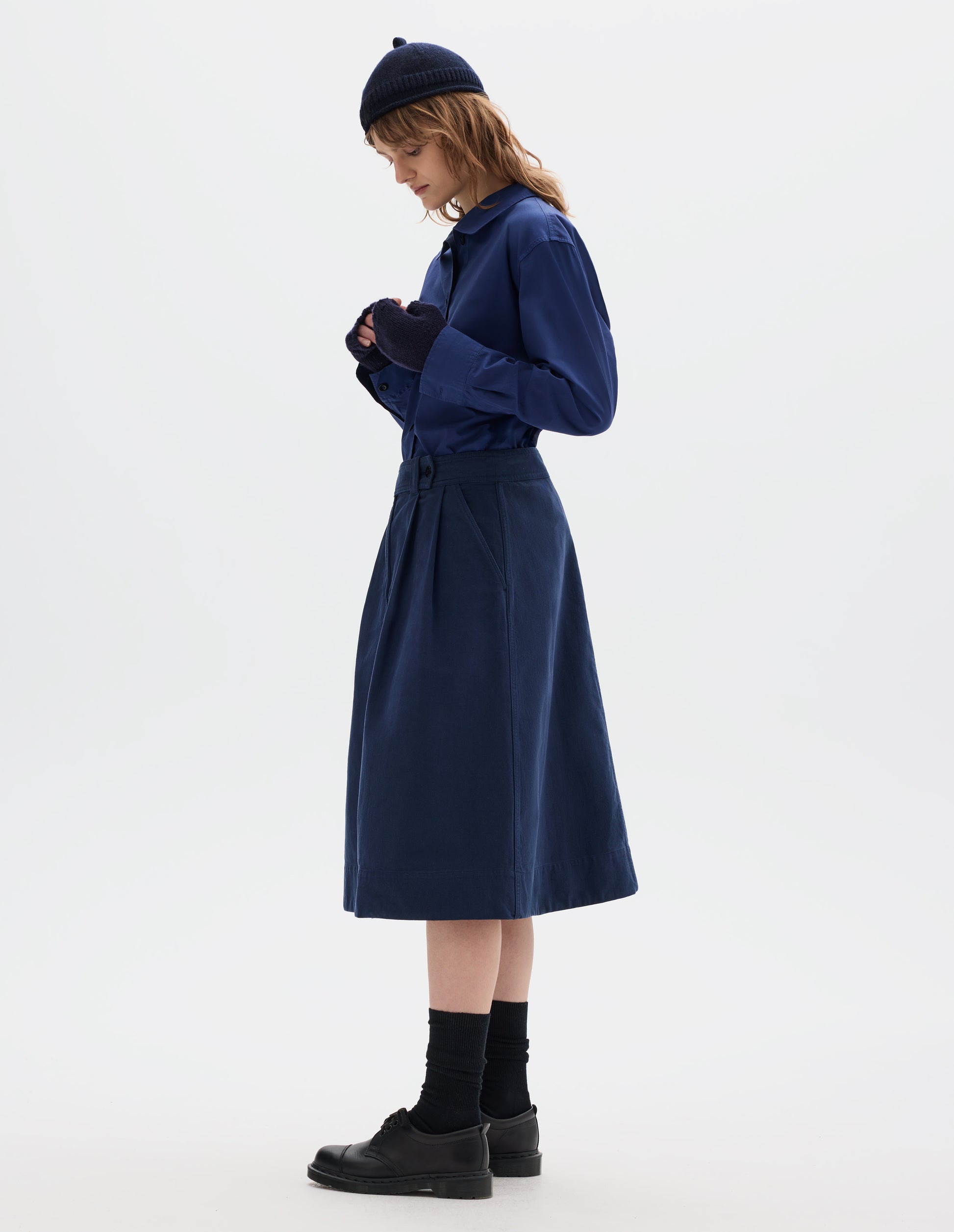 Full body image, side view of model wearing dark blue cotton skirt, with loop and button fastening, slant pockets and ticket pockets. Styled with blue shirt, navy hat and fingerless gloves, black socks and black shoes.