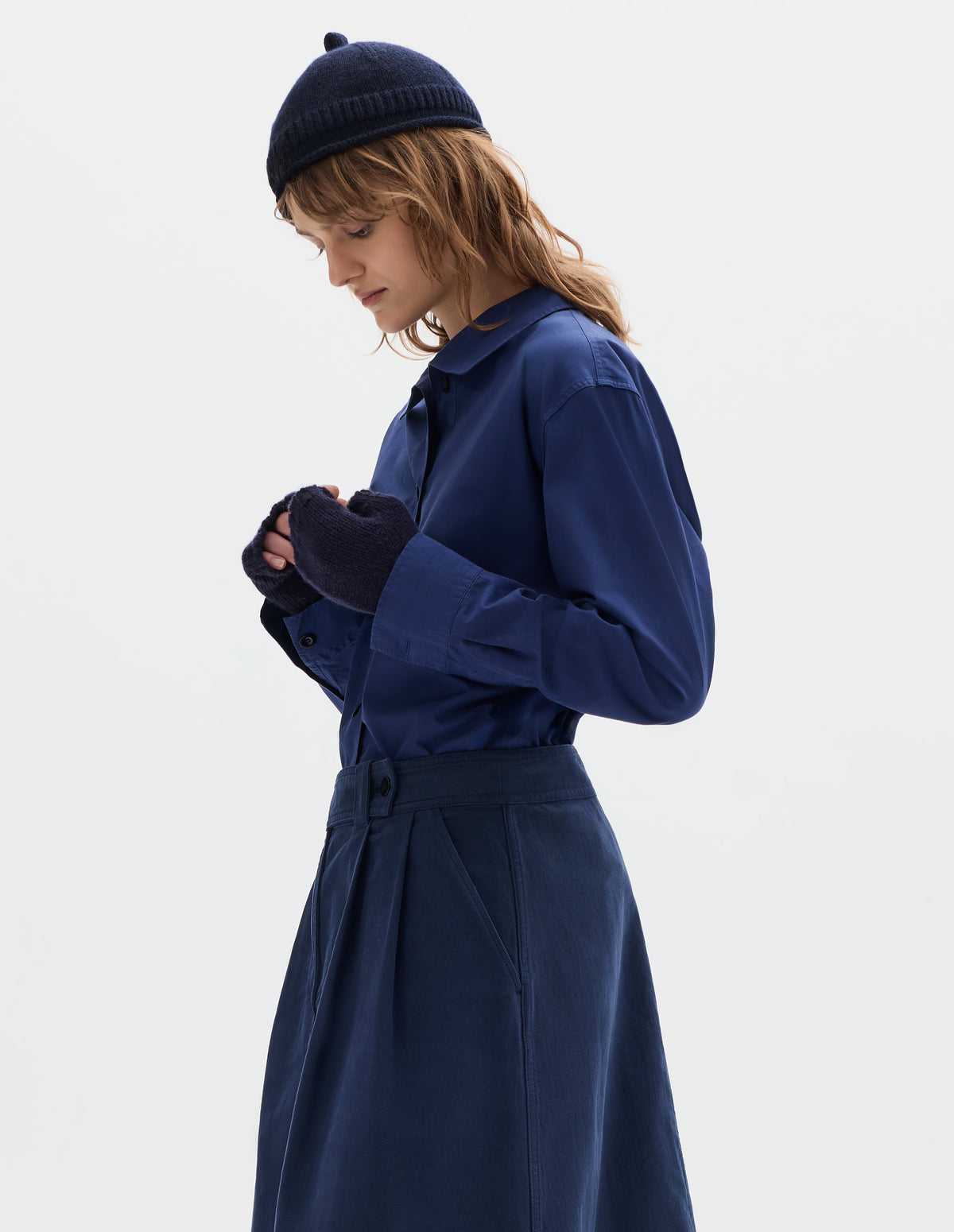 Cropped body image, front view of model wearing a dark blue wool knitted fingerless gloves. Styled with a dark blue beanie with a loop at the top and a rolled hem, blue cotton shirt and pleated skirt.