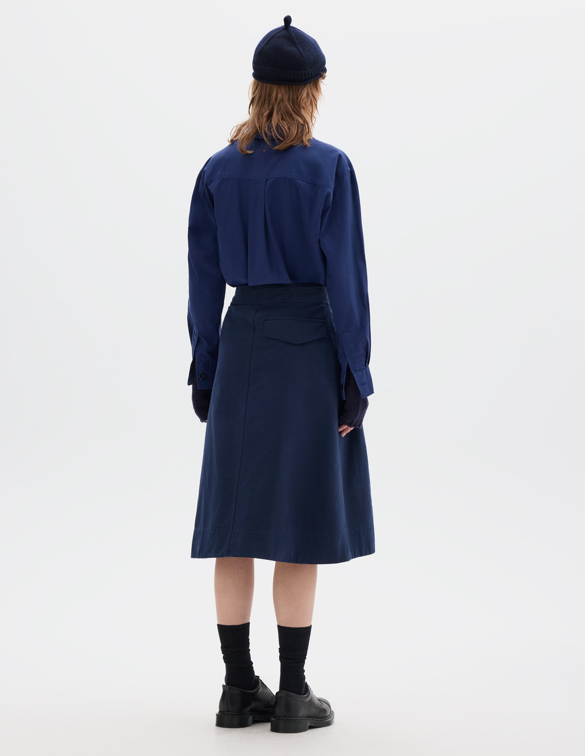 Full body image, back view of model wearing dark blue cotton skirt, with back flap pocket. Styled with blue shirt, navy hat and fingerless gloves, black socks and black shoes.
