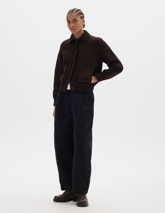 Full body image, front view of model wearing a dark brown boxy fit collared cardigan, with laid-on side entry pockets, and rib detailing on collar, placket, hem and cuffs. Styled with black pleated trousers, brown leather shoes and worn over a white t shirt.