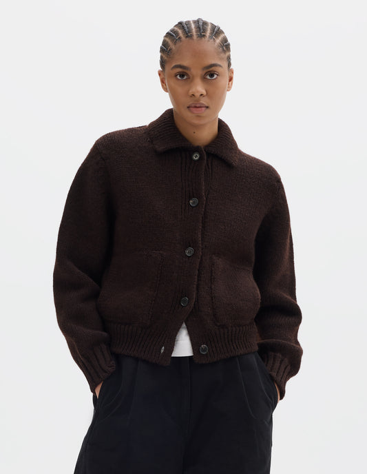Cropped body image, front view of model wearing a dark brown boxy fit collared cardigan, with laid-on side entry pockets, and rib detailing on collar, placket, hem and cuffs. Styled with black pleated trousers and worn over a white t shirt.