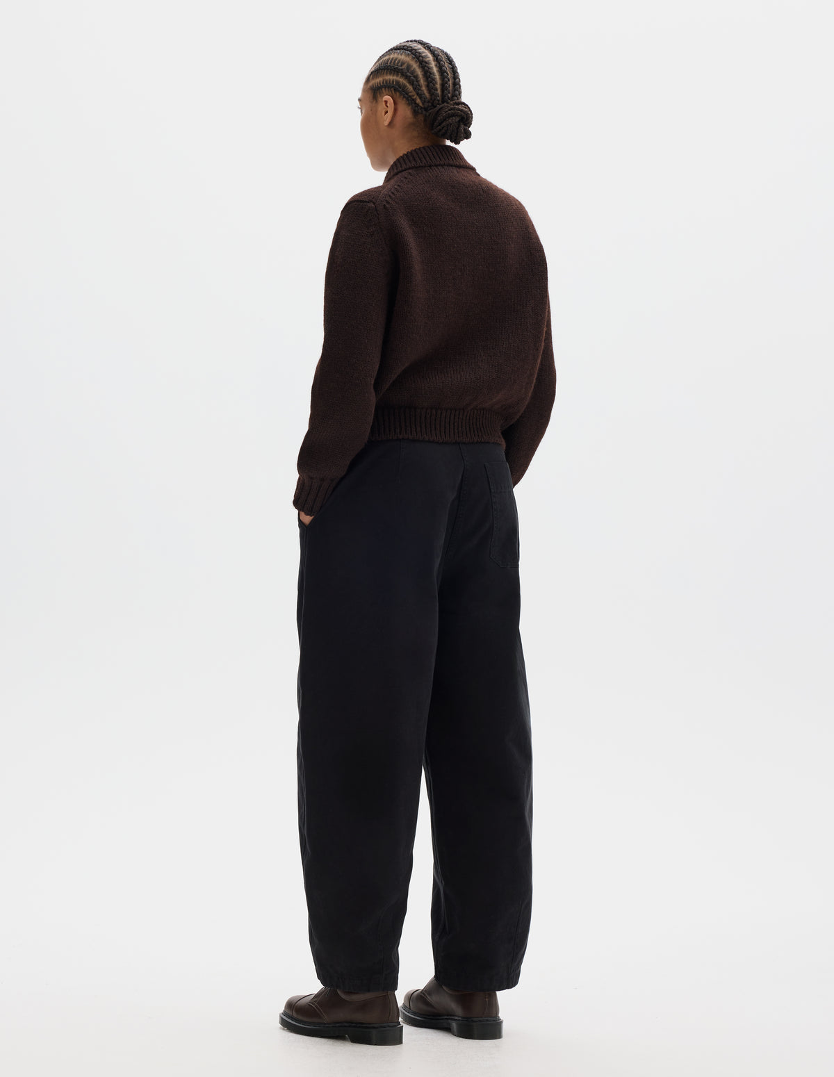 Full body image, back view of model wearing a dark brown boxy fit collared cardigan, rib detailing on collar, hem and cuffs. Styled with black trousers, brown leather shoes.