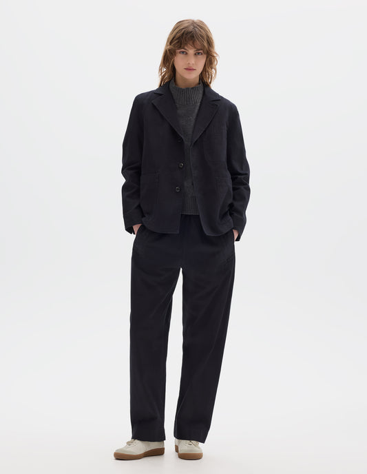 Full body image, front view of model wearing indigo twill single breasted blazer, with two front patch pockets, fold back split cuff, curved hem and corozo button closure. Styled over grey wool slipover with matching indigo twill trousers and white suede trainers.