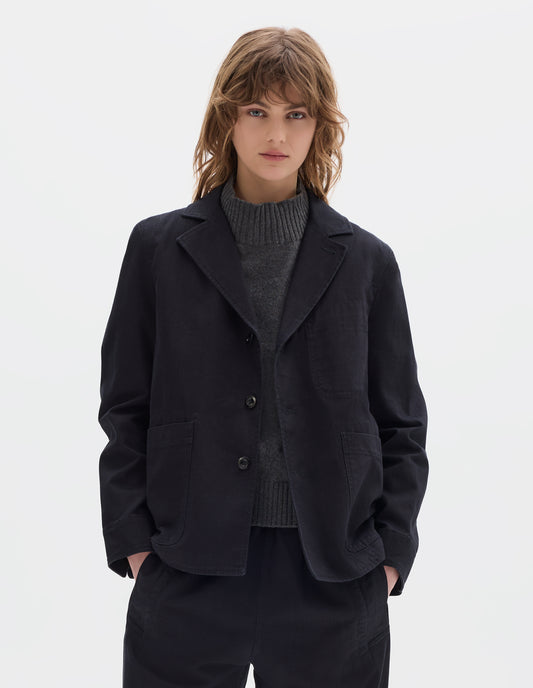 Cropped body image, front view of model wearing indigo twill single breasted blazer, with two front patch pockets, fold back split cuff, curved hem and corozo button closure. Styled over grey wool slipover with matching indigo twill trousers.