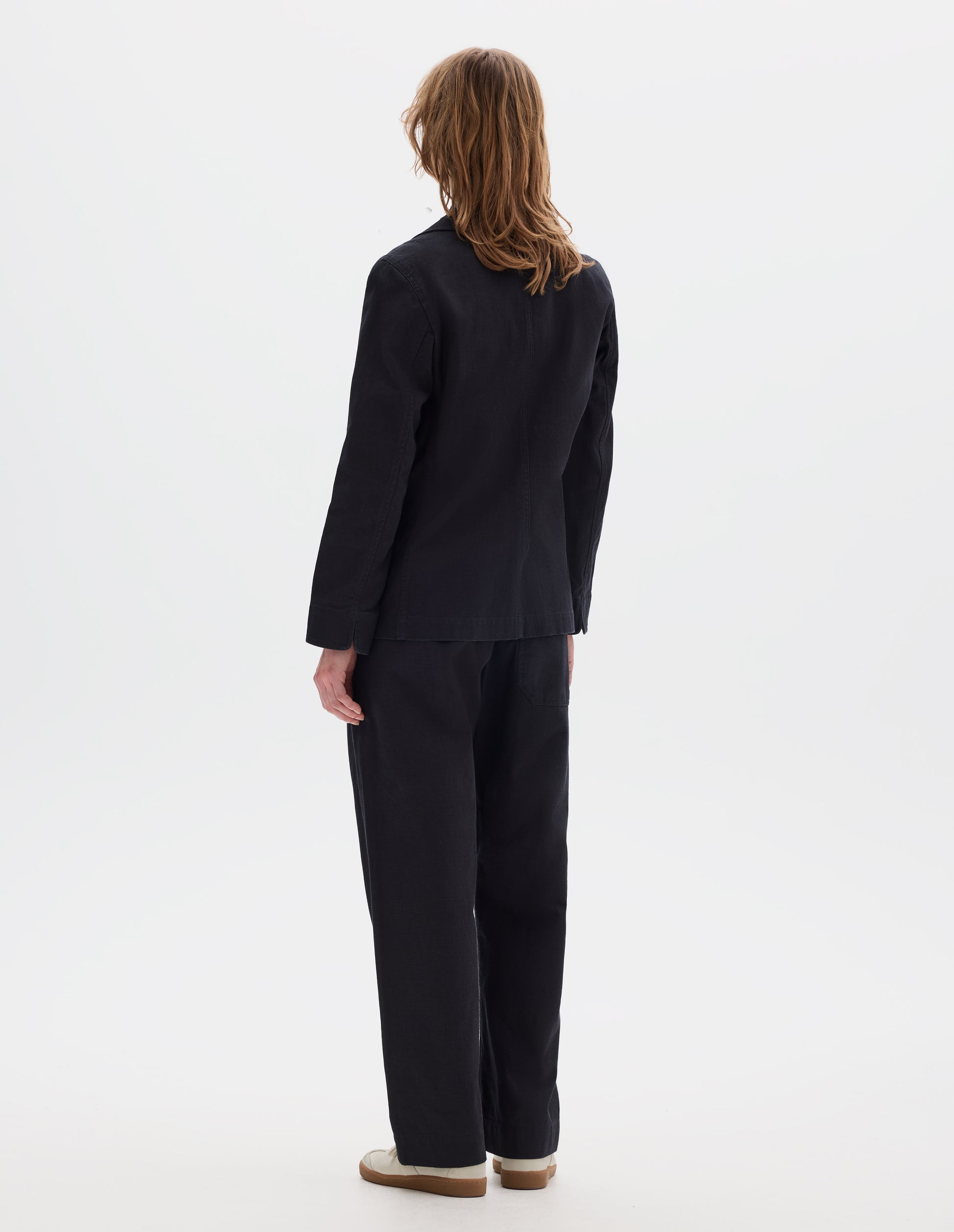 Full body image, back view of model wearing indigo twill single breasted blazer, with fold back split cuff and curved hem. Styled with matching indigo twill trousers and white suede trainers.