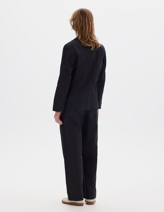 Full body image, back view of model wearing indigo twill single breasted blazer, with fold back split cuff and curved hem. Styled with matching indigo twill trousers and white suede trainers.