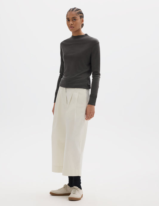 Full body image, front view of model wearing dark grey long sleeve t shirt with set-in sleeves, a rib neck and cuffs with flatlock stitching. Styled with white cropped trousers, black socks and white trainers with gum sole. 
