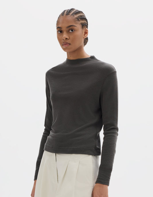 Cropped body image, front view of model wearing dark grey long sleeve t shirt with set-in sleeves, a rib neck and cuffs with flatlock stitching. Styled with white cropped trousers.