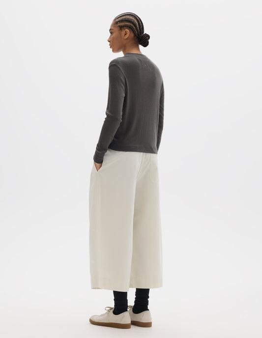 Full body image, back view of model wearing dark grey long sleeve t shirt with set-in sleeves, a rib neck and cuffs with flatlock stitching. Styled with white cropped trousers, black socks and white trainers with gum sole. 