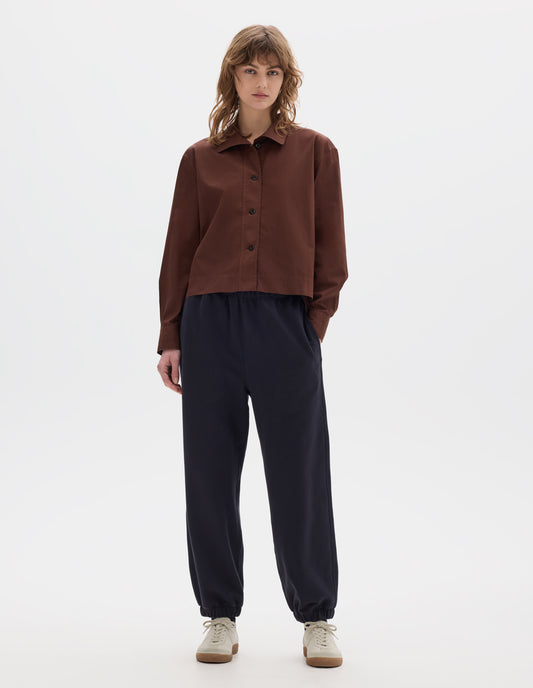 Full body image, front view of model wearing a reddish brown cotton cropped shirt with asymmetric collar with a softly rounded point. Styled with dark navy jogger style trousers with elasticated waistband and hem and off white suede trainers.
