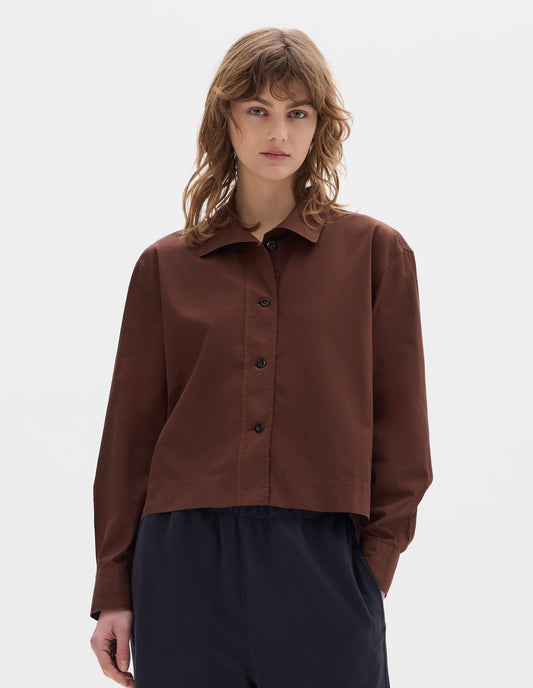 Cropped body image, front view of model wearing a reddish brown cotton cropped shirt with asymmetric collar with a softly rounded point. Styled with dark navy jogger style trousers.