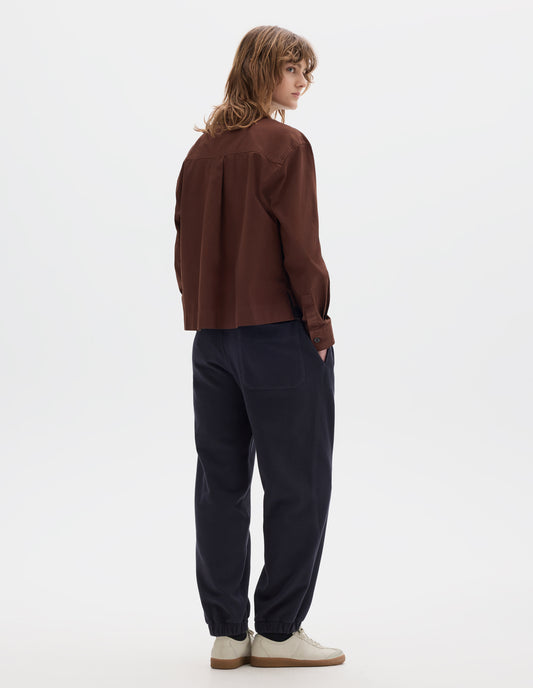 Full body image, back view of model wearing a reddish brown cotton cropped shirt with a box pleat on back yoke. Styled with dark navy jogger style trousers with elasticated waistband and hem and off white suede trainers.